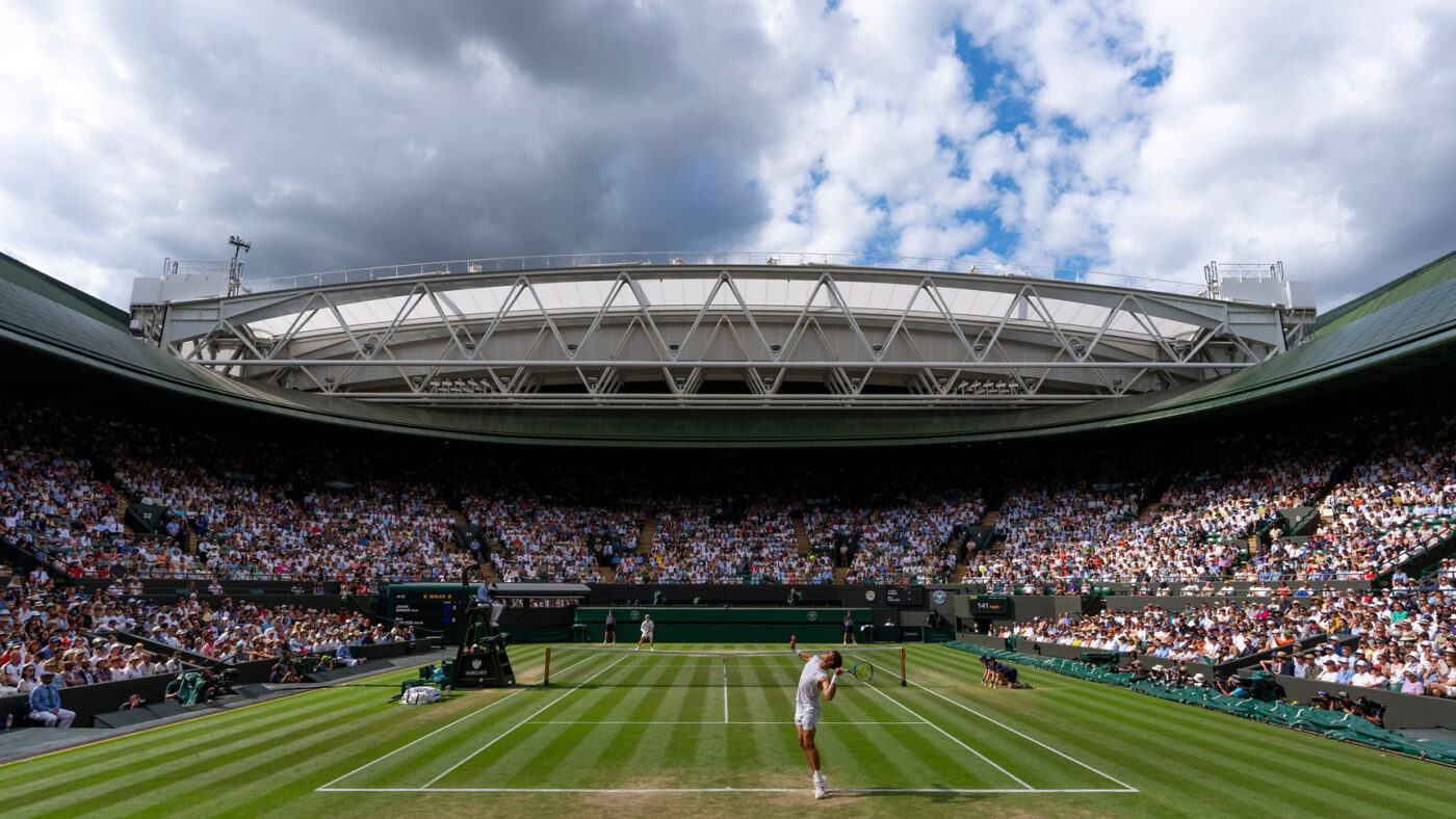 A men's singles match in progress on Centre Court at Wimbledon, with the retractable roof open above a packed crowd